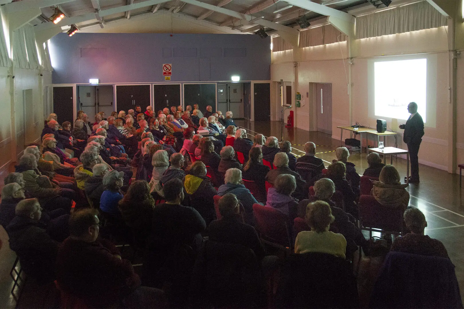 Audience of people seated in chairs watching a presentation. A speaker stands at a projector screen.