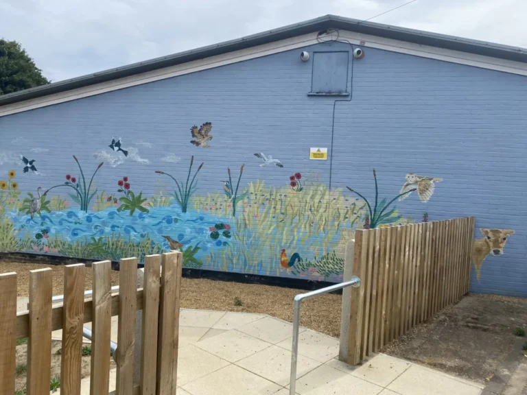 A mural of a pond scene with birds, flowers, and plants is painted on the side of a light blue brick building behind a wooden fence.