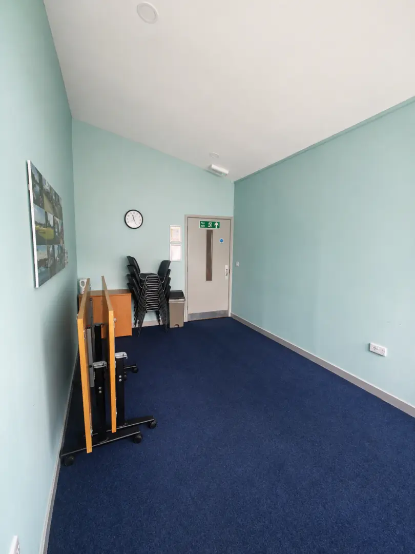 Small conference room with blue carpet, light blue walls, a door, chairs stacked in the corner, a wall clock, and a wooden lectern. Emergency exit sign is above the door.