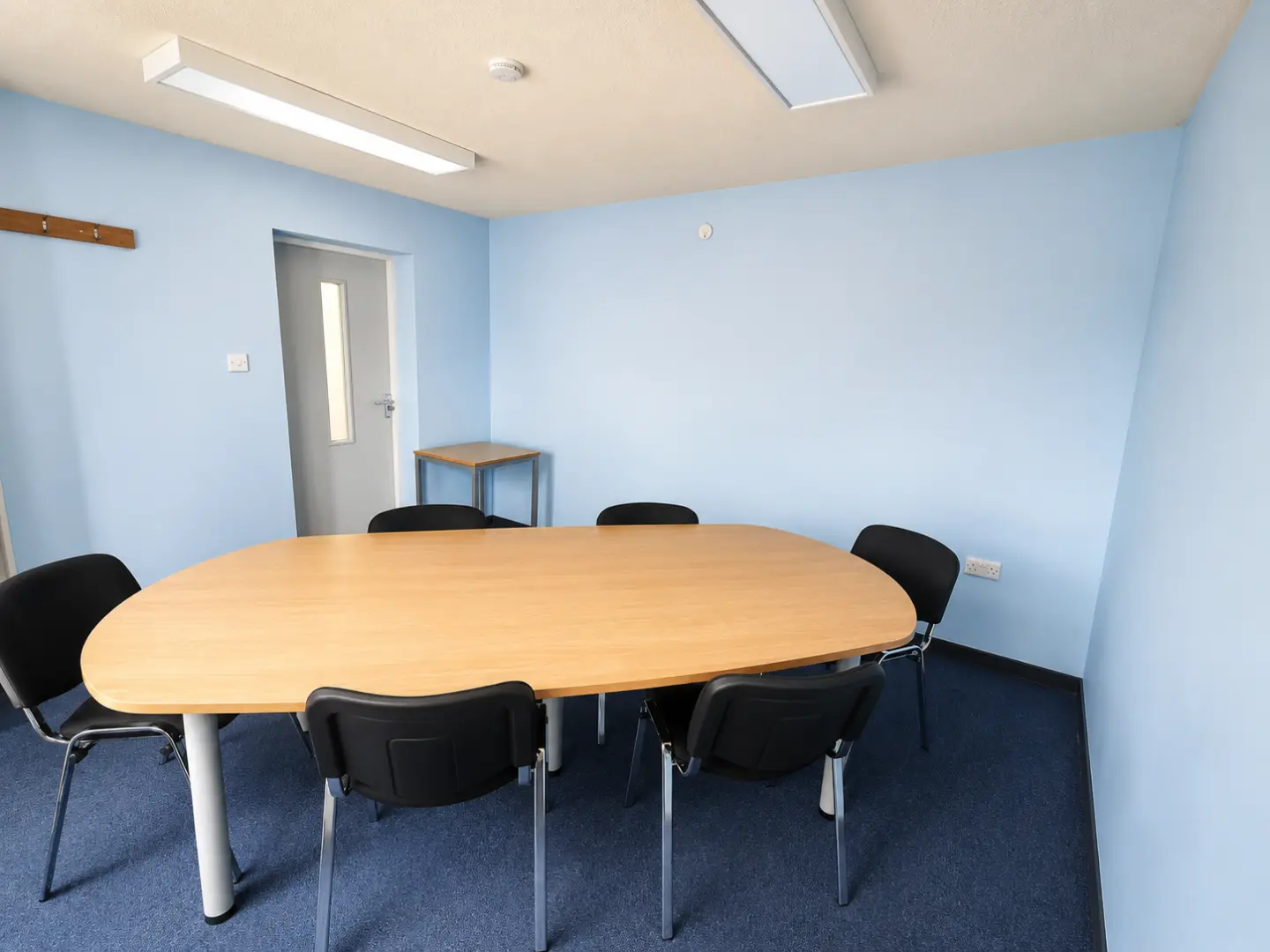 A small meeting room with a wooden oval table, six black chairs, light blue walls, blue carpet, and a door with a window.